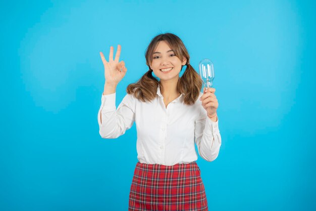 Una hermosa colegiala sonriente sosteniendo una bombilla de idea y gesticulando OK. foto de alta calidad