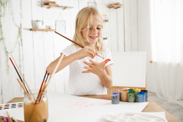 Hermosa, creativa y ocupada niña rubia en camiseta blanca dibujando en su palma con un pincel.