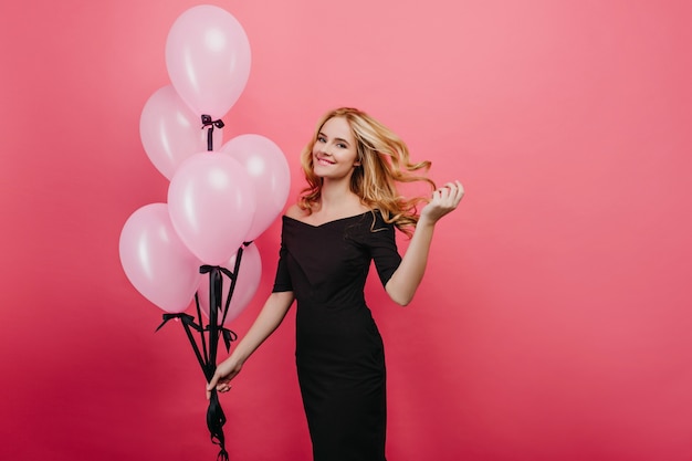 Hermosa cumpleañera juega con su cabello claro. Foto interior de mujer rubia atractiva en vestido largo posando con globos de helio.
