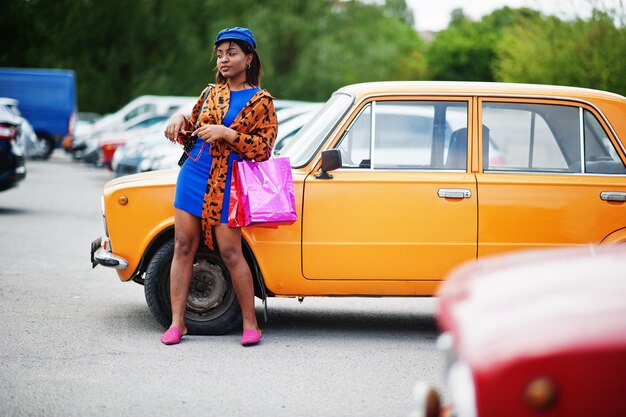 Hermosa dama afroamericana con bolsas de compras de pie cerca de un auto retro clásico naranja