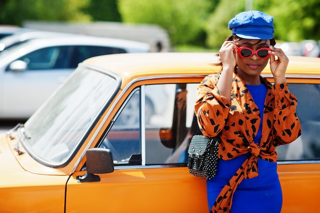 Hermosa dama afroamericana con gafas de sol de pie cerca de un auto retro clásico naranja