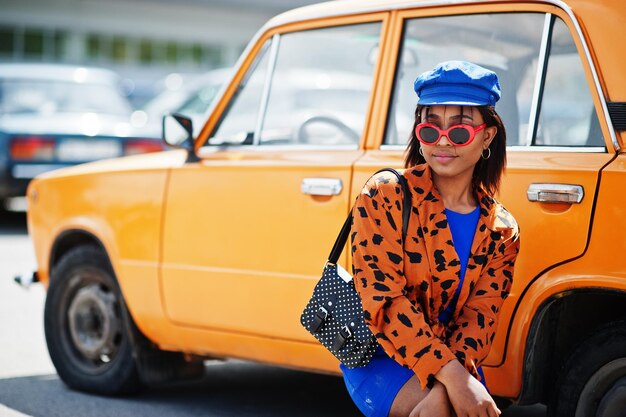 Hermosa dama afroamericana con gafas de sol de pie cerca de un auto retro clásico naranja