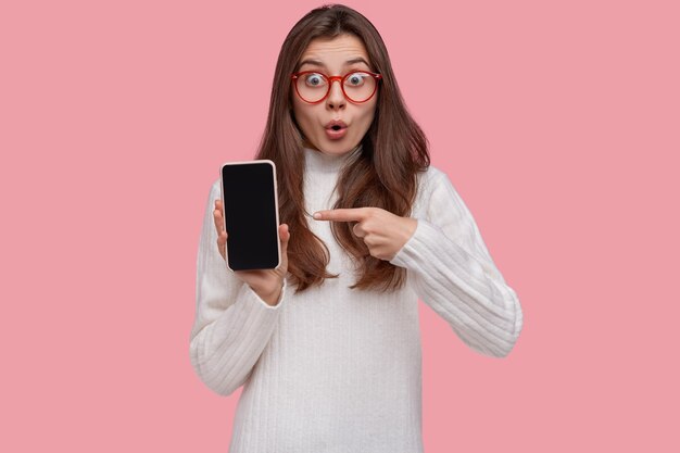 La hermosa dama aturdida apunta al teléfono inteligente con mirada de sorpresa, muestra el dispositivo, usa lentes transparentes y un jersey blanco