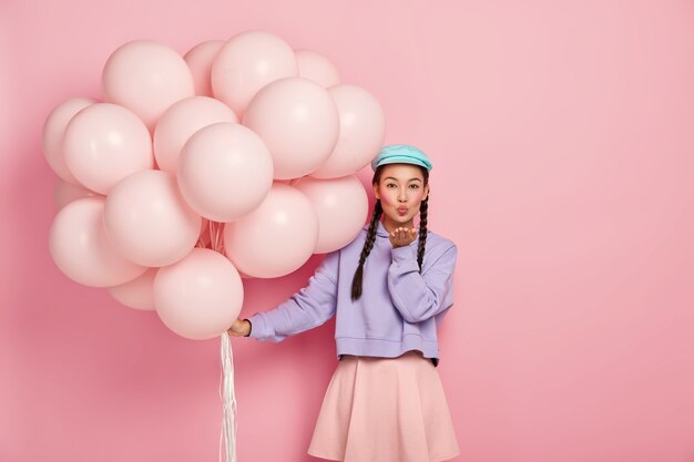 Hermosa dama de cabello oscuro con apariencia específica, usa maquillaje, mantiene los labios redondeados, sopla beso de aire a la cámara, tiene expresión coqueta, posa con globos de helio, aislado sobre una pared rosa