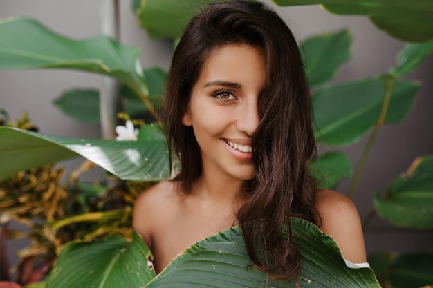 Hermosa dama con cabello rizado y ojos verdes sonríe dulcemente Mujer con pestañas largas posando contra el telón de fondo de una planta tropical