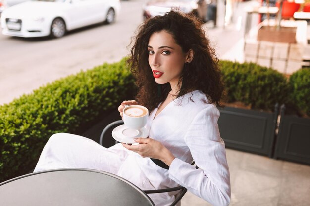 Hermosa dama con cabello rizado oscuro en traje blanco sentada en la mesa con una taza de café en las manos y mirando cuidadosamente a la cámara en un café en la calle