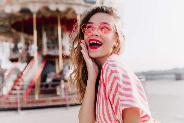 Hermosa dama despreocupada sonriendo en el fondo de la calle y tocando su mejilla Modelo femenino romántico en gafas de sol rosas de pie en el parque de atracciones