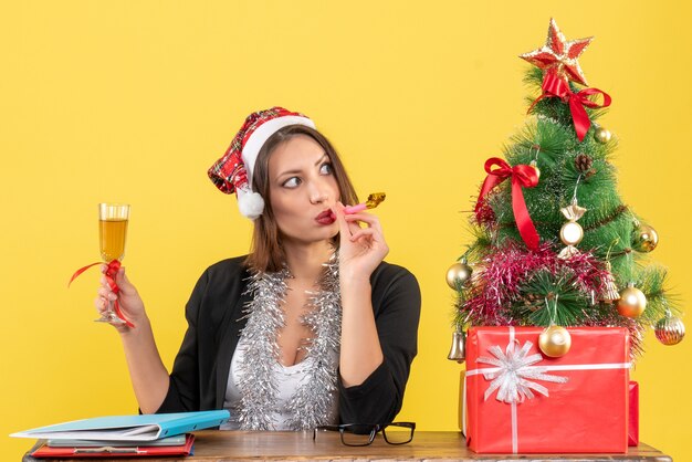 Hermosa dama encantadora en traje con sombrero de santa claus y adornos de año nuevo levantando vino en la oficina en amarillo aislado