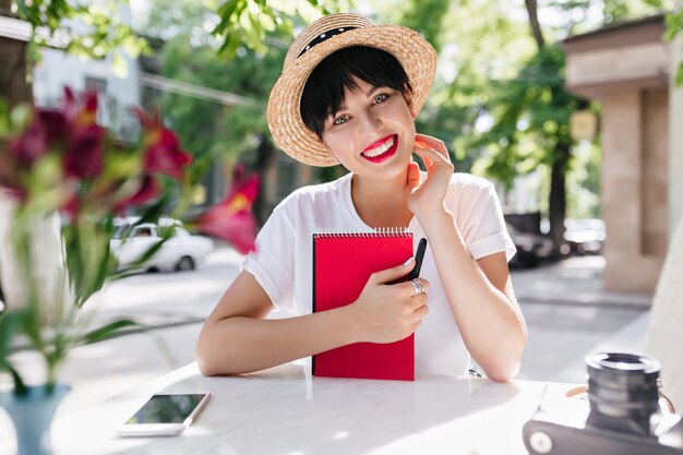 Hermosa dama con expresión de cara feliz con cuaderno rojo descansando en la cafetería al aire libre con teléfono