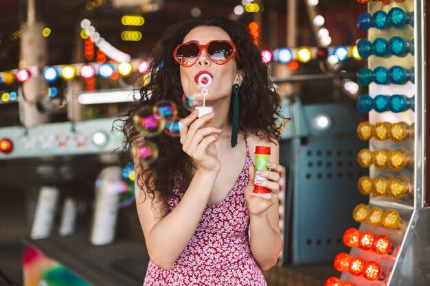 Hermosa dama con gafas de sol de corazón y vestido de pie y soplando burbujas mientras pasa tiempo en el parque de diversiones