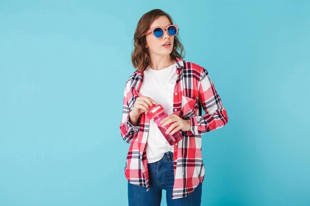 Hermosa dama con gafas de sol de pie con una botella deportiva rosa sobre fondo azul Foto de una linda chica hipster con camisa a cuadros y jeans