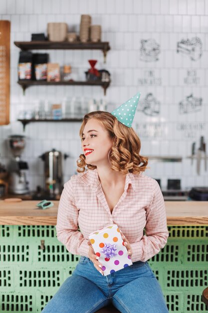 Hermosa dama con gorra de cumpleaños sentada en el mostrador del bar con una caja de regalo en las manos y felizmente mirando a un lado en el café