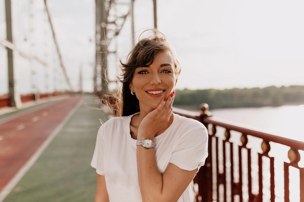 Hermosa dama con grandes ojos azules sonriendo a la cámara y tocando la cara mientras camina por la ciudad por la mañana