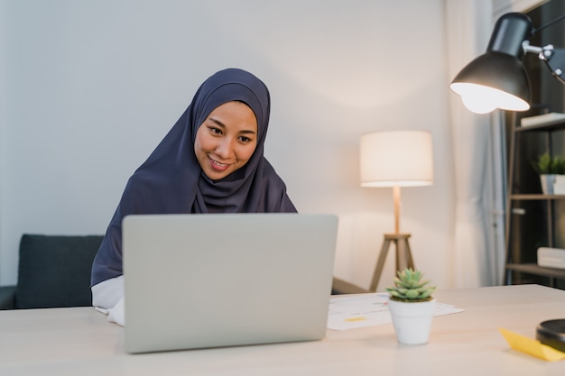 Hermosa dama musulmana de Asia en ropa casual con pañuelo en la cabeza usando la computadora portátil en la sala de estar en la casa de noche.