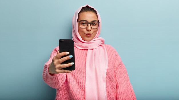 Hermosa dama musulmana de cabello oscuro graba videos, mantiene los labios doblados, se toma una selfie, captura una nueva perspectiva, posa con una bufanda rosa y un suéter, publica fotos en línea para los seguidores, hace una toma genial en interiores