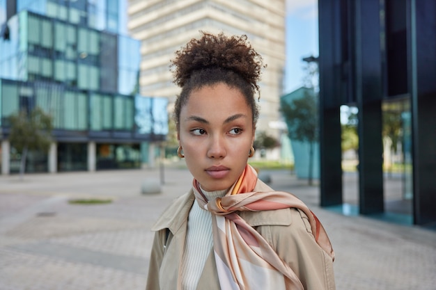 Hermosa dama de piel oscura con cabello rizado camina en un entorno urbano enfocado en algún lugar usa un pañuelo impermeable atado sobre el cuello tiene una expresión contemplativa va a trabajar poses en el área de publicidad