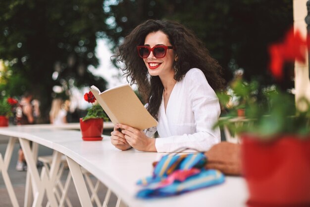 Hermosa dama sonriente con cabello oscuro y rizado con gafas de sol de pie con un bloc de notas en las manos mientras pasa felizmente el tiempo en la terraza de verano de la cafetería