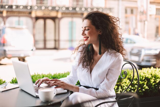Hermosa dama sonriente con cabello rizado oscuro en traje blanco sentada en la mesa con una taza de café y trabajando en una computadora portátil mientras pasa tiempo en un café en la calle con vista a la ciudad en el fondo