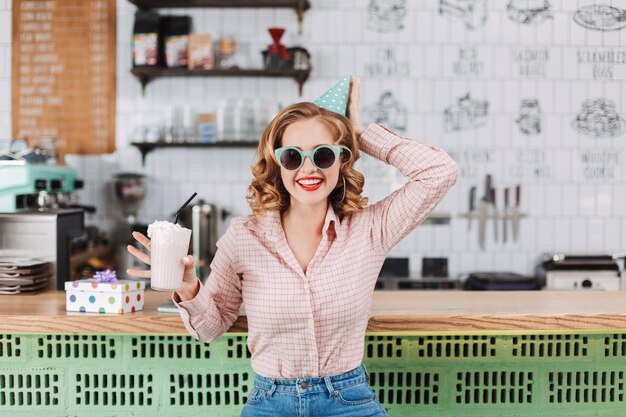 Hermosa dama sonriente con gafas de sol y gorra de cumpleaños sentada en el mostrador del bar con un batido en la mano y felizmente mirando a la cámara en el café