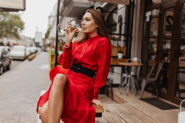 Hermosa dama con vestido de diseñador caro bebe delicioso vino espumoso de cristal. Foto de cuerpo entero de blogger sentado en la cafetería