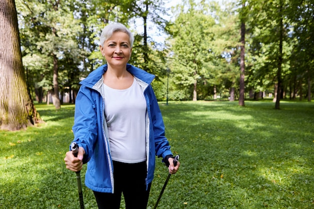 Hermosa deportista madura disfrutando de su pasatiempo activo, sosteniendo bastones para caminar escandinavo, teniendo cuerpo en forma, pasando los días de jubilación en actividades saludables. Verano, deporte y ocio