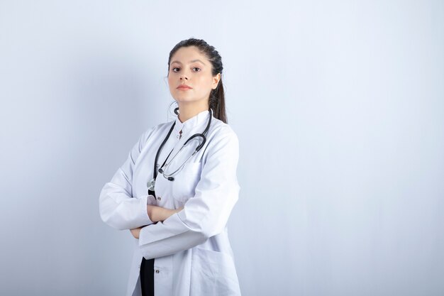 Hermosa doctora en bata blanca con los brazos cruzados sobre la pared blanca.