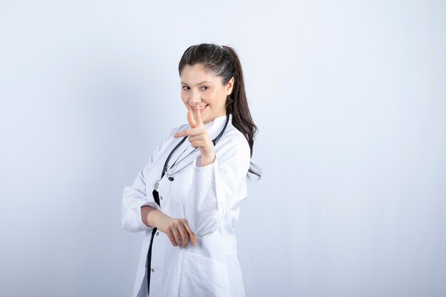 Hermosa doctora en bata blanca posando con estetoscopio sobre pared blanca.