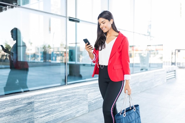 Hermosa ejecutiva sonriente escuchando música a través de auriculares mientras usa el teléfono móvil fuera de la oficina