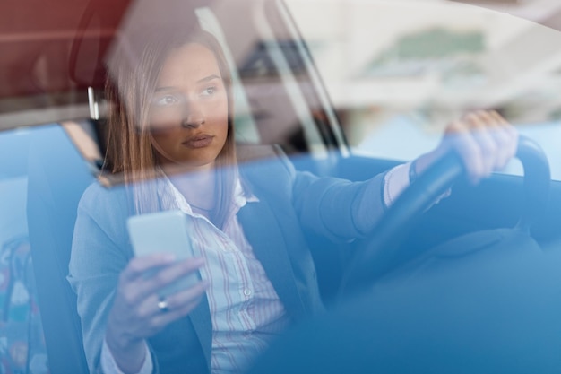 Hermosa empresaria conduciendo al trabajo y usando el teléfono celular detrás del volante La vista es a través del cristal