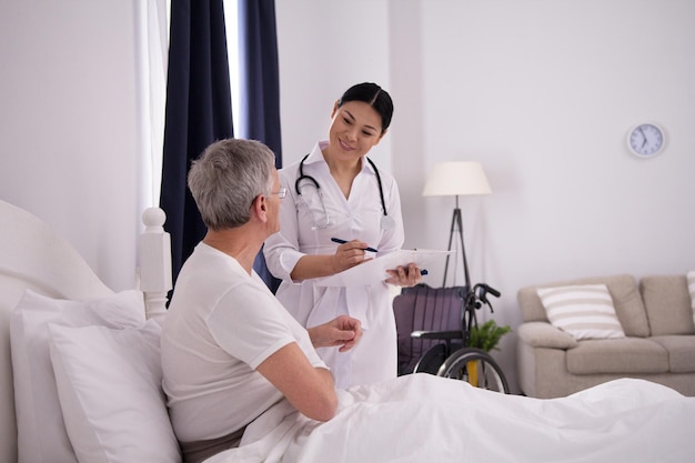 Hermosa enfermera haciendo un chequeo rápido al paciente. Alegre y amigable médico asiático sosteniendo el portapapeles para verificar cómo le está yendo a su anciano paciente.