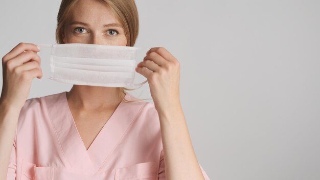 Hermosa enfermera rubia con máscara médica en cámara con lugar para publicidad o texto promocional cerca de fondo blanco