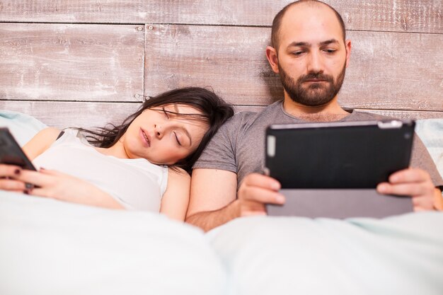 Hermosa esposa en pijama durmiendo mientras su esposo está trabajando en una tableta.