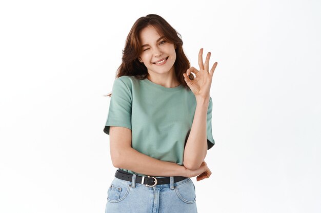 Hermosa estudiante adolescente, sonriendo y mostrando un signo bien, le gusta el artículo, aprueba y elogia la buena elección, garantiza la calidad, recomienda el producto, de pie contra la pared blanca