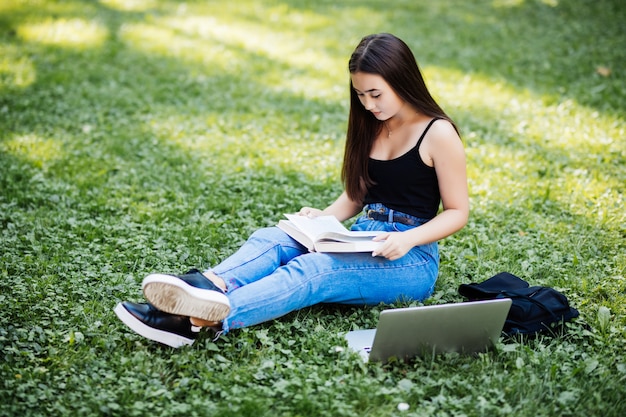 Hermosa estudiante asiática manteniendo registros en un cuaderno de papel mientras estudia en línea con una computadora portátil, sentado en el césped, al aire libre