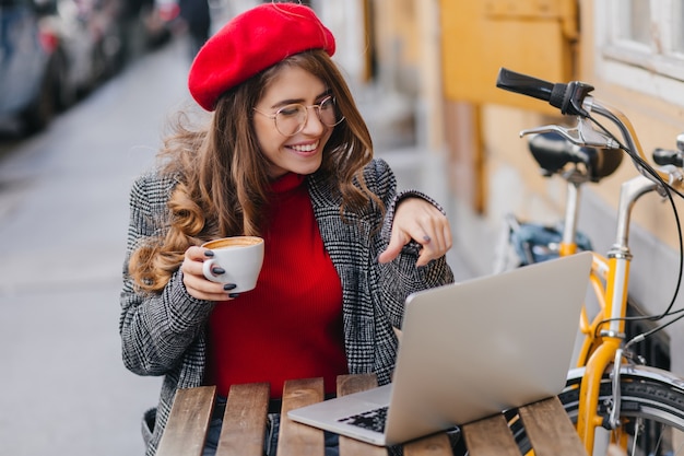 Hermosa estudiante de boina roja tomando café mientras trabaja con la computadora