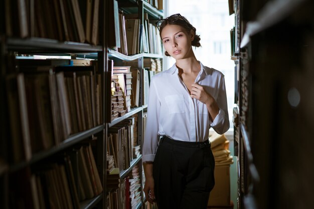 Hermosa estudiante con una camisa blanca se encuentra entre las filas de la biblioteca