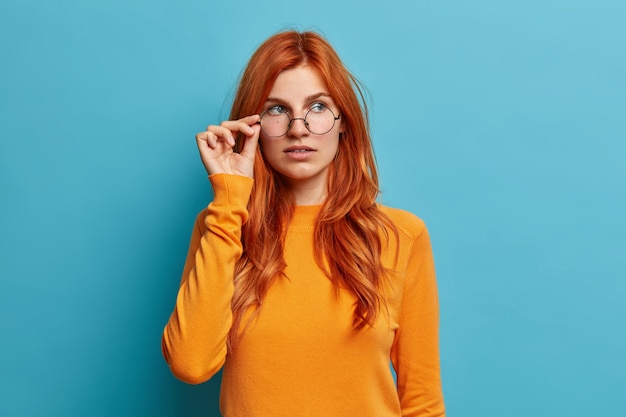 Hermosa estudiante de jengibre piensa en un proyecto futuro y mantiene la mano en las gafas concentradas pensativamente a un lado vestida con un jersey naranja casual.