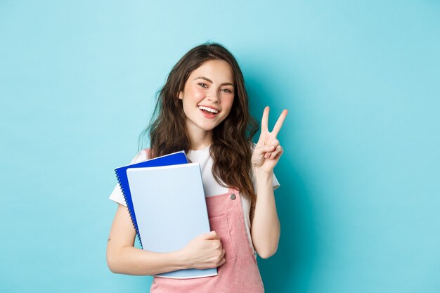 Hermosa estudiante mostrando v-sign y sonriendo feliz, sosteniendo cuadernos con material de estudio, asistiendo a cursos, de pie sobre fondo azul.