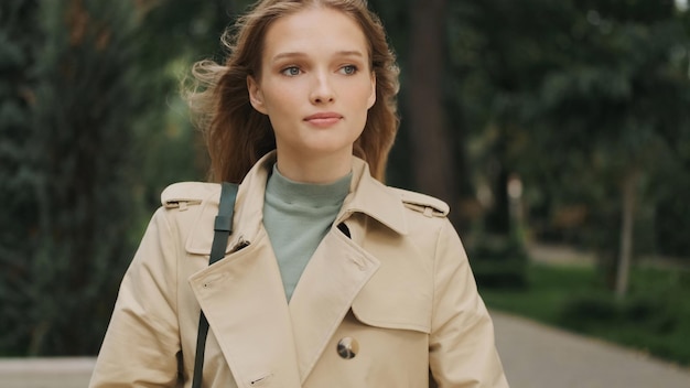 Hermosa estudiante pensativa vestida con una gabardina que va a la universidad a través del parque de la ciudad y se viste con algo