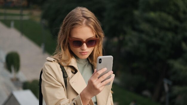 Hermosa estudiante rubia con gafas de sol que se ve confiada tomando selfie en el teléfono inteligente en el parque de la ciudad Tecnología moderna