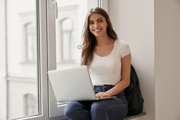Hermosa estudiante sentada con una laptop en el alféizar de la ventana