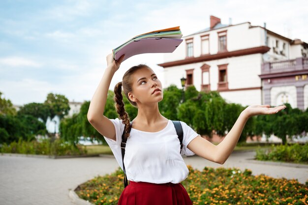 Hermosa estudiante sosteniendo carpetas por encima de la cabeza, escondiéndose de la lluvia.