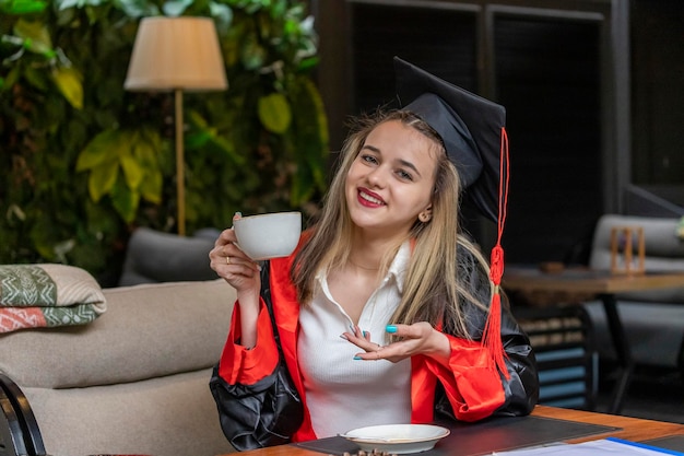 Hermosa estudiante viste capa de graduación y bebe café en el restaurante