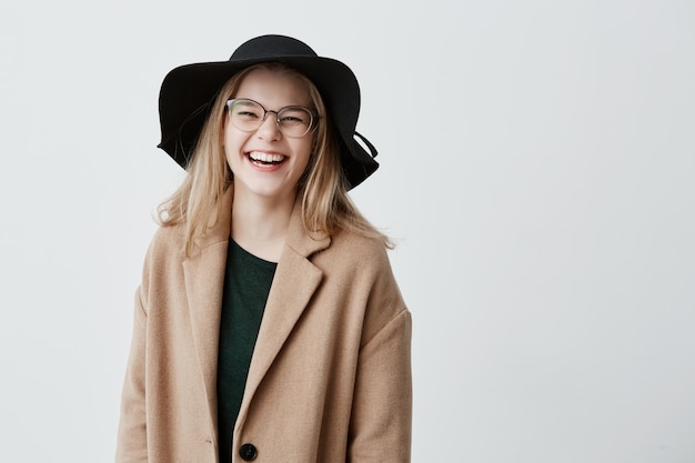 Hermosa eufórica adolescente con elegantes lentes, sombrero negro y abrigo, disfrutando de su vida, mirando y sonriendo, llena de alegría y felicidad. Concepto de personas y estilo de vida