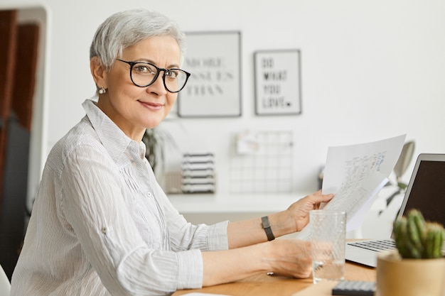 Hermosa exitosa empresaria madura confiada con cabello gris corto trabajando en su oficina, usando una computadora portátil con papeles en sus manos, estudiando el informe financiero, sonriendo