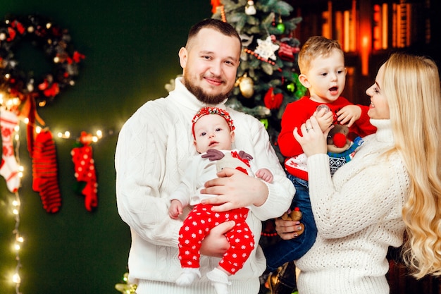 Hermosa familia con niños en cálidos suéteres posa ante una pared verde y rico árbol de Navidad