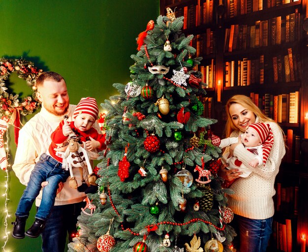 Hermosa familia con niños en cálidos suéteres posa ante una pared verde y rico árbol de Navidad