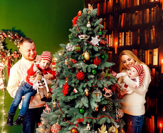 Hermosa familia con niños en cálidos suéteres posa ante una pared verde y rico árbol de Navidad