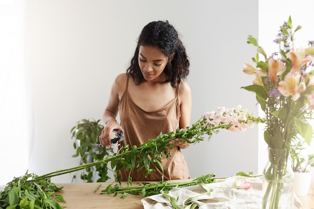 Hermosa floristería femenina africana sonriente cortando tallos trabajando con flores sobre la pared blanca.