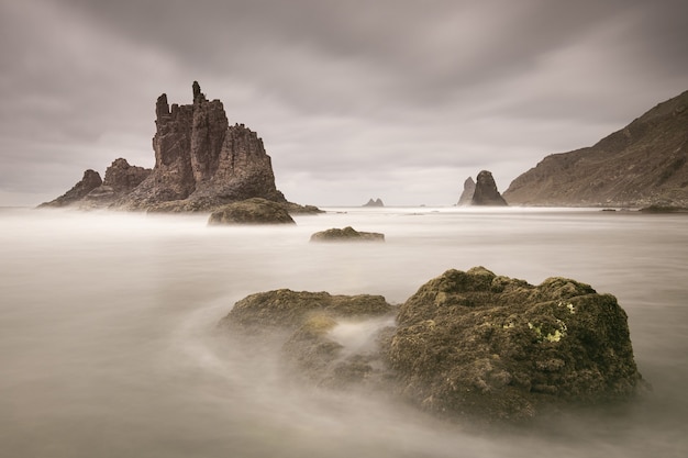 Hermosa foto de agua que fluye alrededor de grandes piedras cerca de la roca de Benijo en un día nublado en España